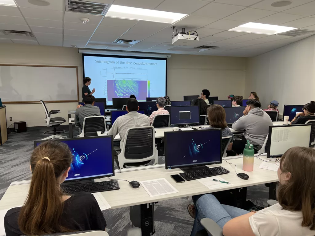 Students sit at rows of computers as an alumni mentor presents a PowerPoint lecture.