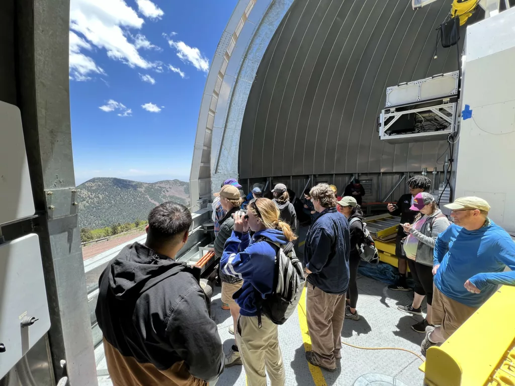 The dome of a large telescope is open, and student and staff look out at the sweeping mountain landscape.