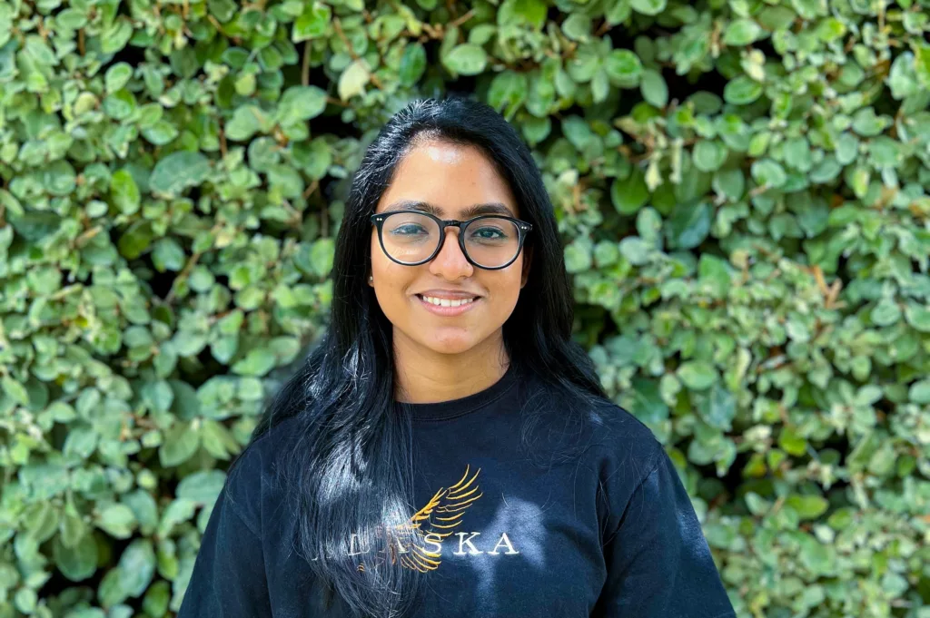 Ann Mariya wearing a black shirt and glasses standing in front of vegetation smiling.