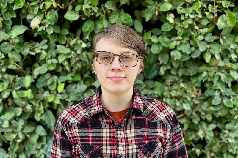 Elliz smiling in front of green foilage wearing a red and black plaid flannel and glasses.