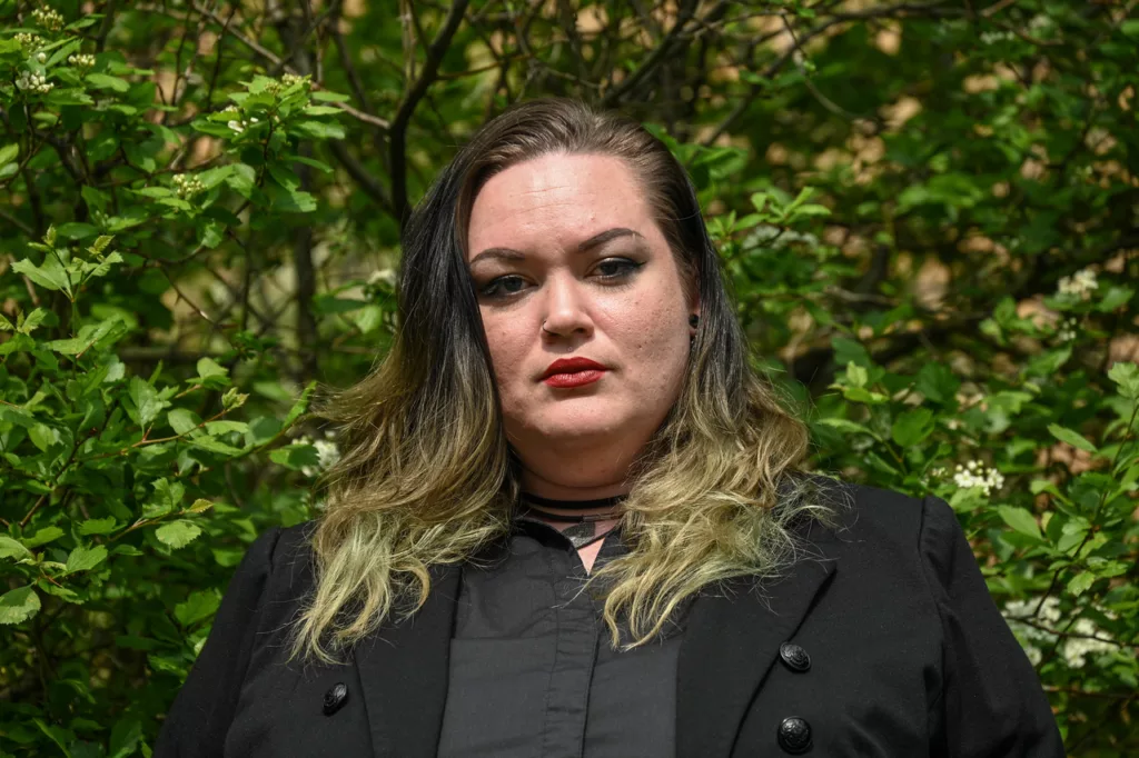 A headshot of Danielle wearing a grey collared shirt underneath a black blazer.