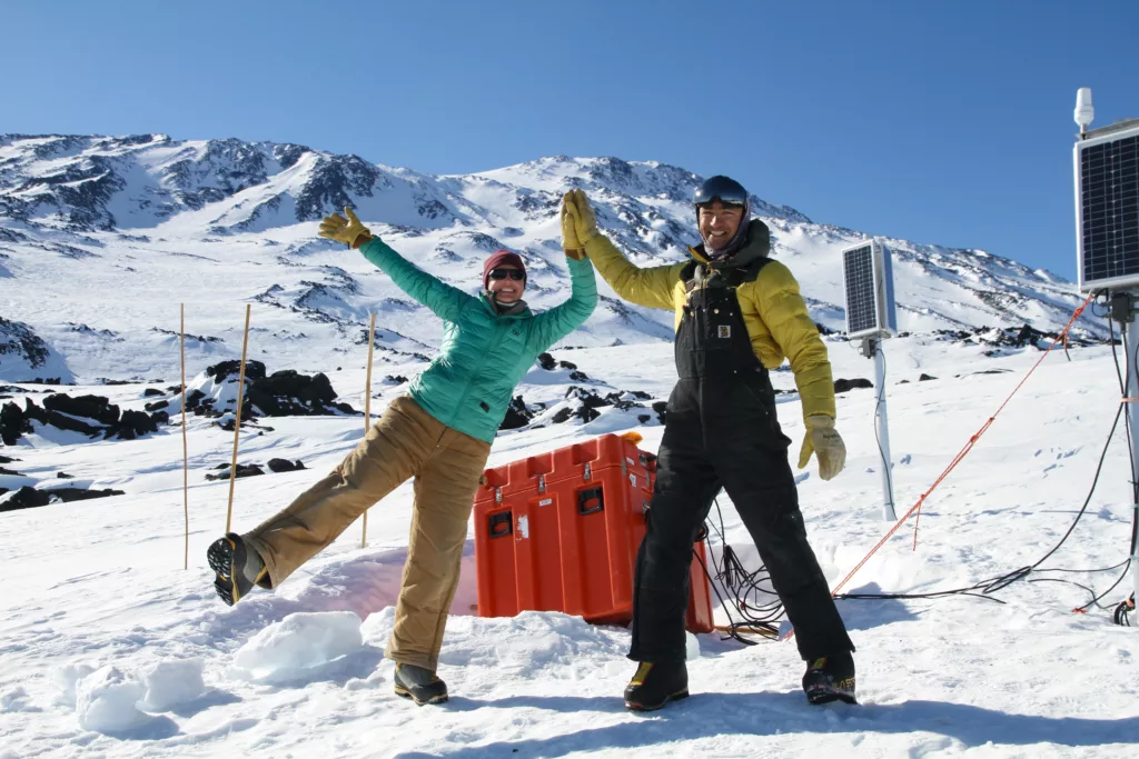 two engineers high-five in front of an equipment case and solar panels in the snow