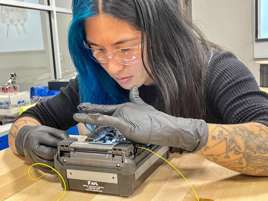 a woman wearing safety glasses and latex gloves manipulates a small device to spice fiber optic cables