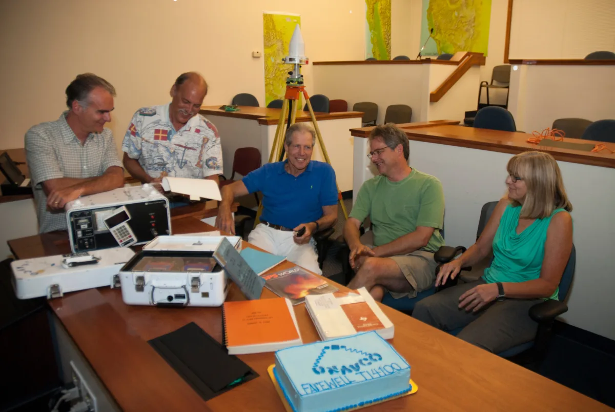 Six people sit and stand around a table during a farewell event for UNAVCO’s first GPS receiver, the TI-4100, on July 25, 2014. Vintage GPS equipment and technical manuals are spread across the table, along with a decorated cake that reads “UNAVCO Farewell TI-4100.” A GPS antenna setup is visible in the background, with topographic maps on the walls.