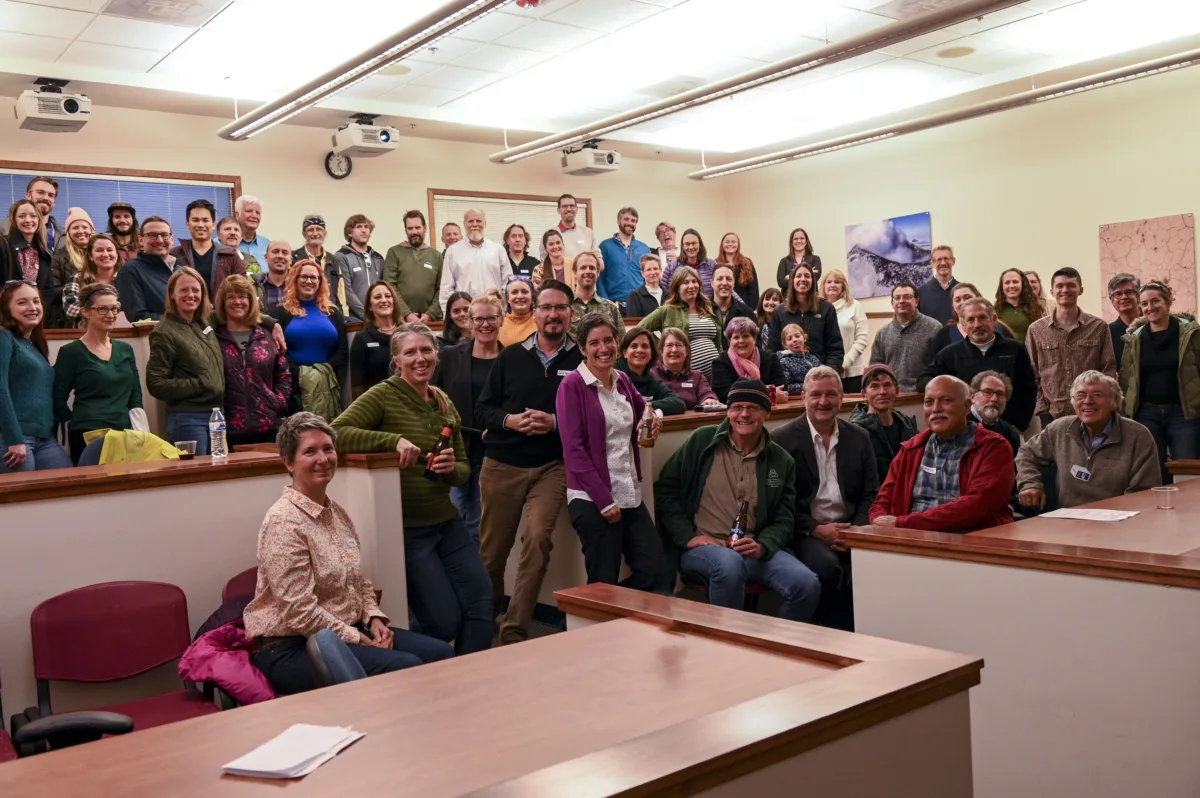 A large group of UNAVCO staff and community members gather for a celebratory group photo at the UNAVCO office in Boulder, Colorado, in October 2022. Smiling participants fill a tiered classroom space, some holding drinks, marking the organization’s legacy ahead of its merger into EarthScope Consortium.