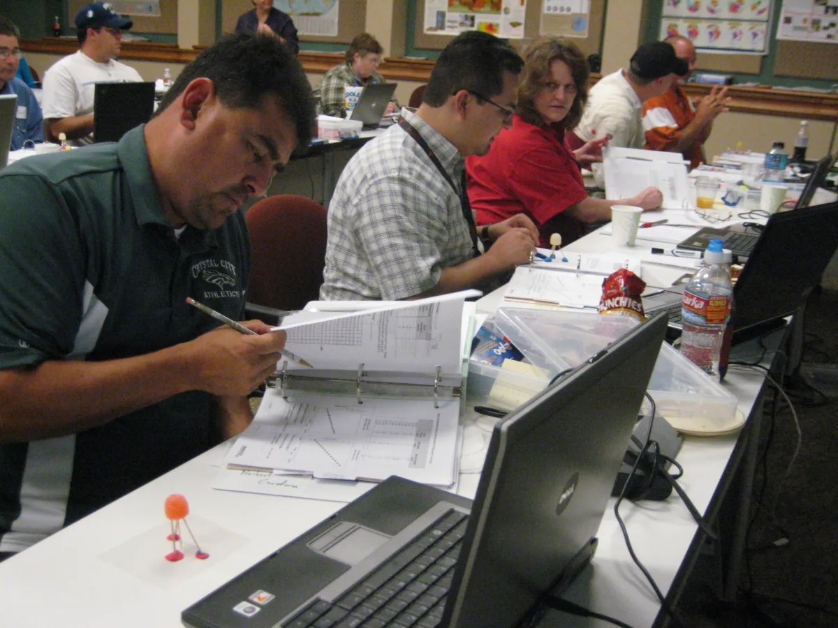 Participants in a UNAVCO Education & Outreach (E&O) short course sit at a long table working with laptops and printed materials. In the foreground, a participant examines a binder while a small gumdrop-and-toothpick model of a GPS station—nicknamed “gumdrop GPS”—sits nearby. Other attendees engage with data sheets and models, illustrating hands-on learning about geodetic motion.