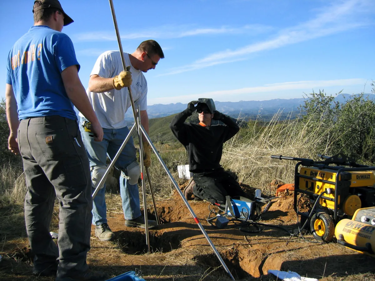 Three field engineers install the first Plate Boundary Observatory (PBO) GPS station at P295 Chimney Rock Ranch in Lake Nacimiento, California, on January 9, 2004. One person kneels beside a power drill while another steadies a metal tripod over a hole. A third person stands nearby wearing a UNAVCO shirt. Tools and a yellow generator are visible, with rolling hills and blue sky in the background.