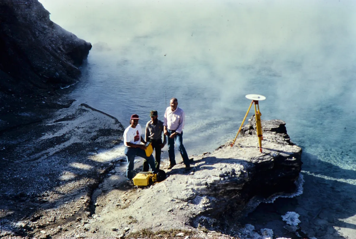 Three geoscientists conduct a GPS survey beside a steaming hydrothermal pool at Yellowstone National Park in 1989. A tripod-mounted GPS antenna is set up on a rocky outcrop. From left to right: Chuck Meertens, Rick Hutchinson, and Bob Smith stand near yellow equipment cases at the water’s edge.