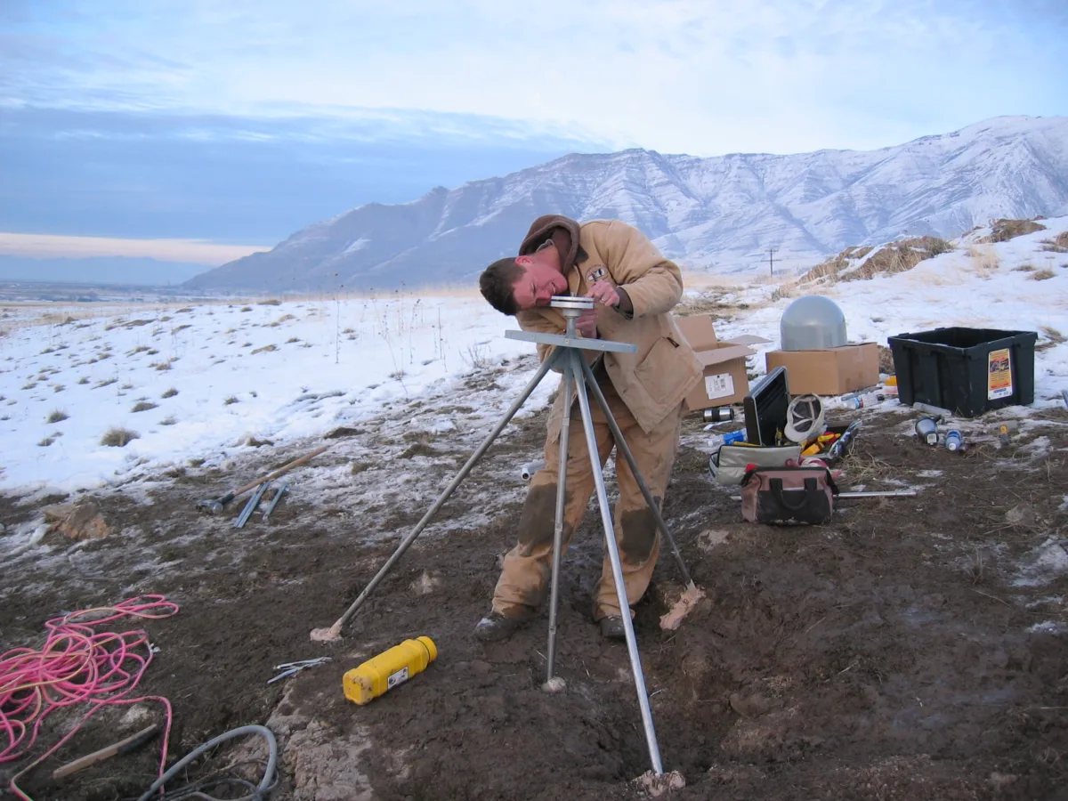 A worker adjusts the base of a GPS antenna, with snow and a ridgeline in the background.