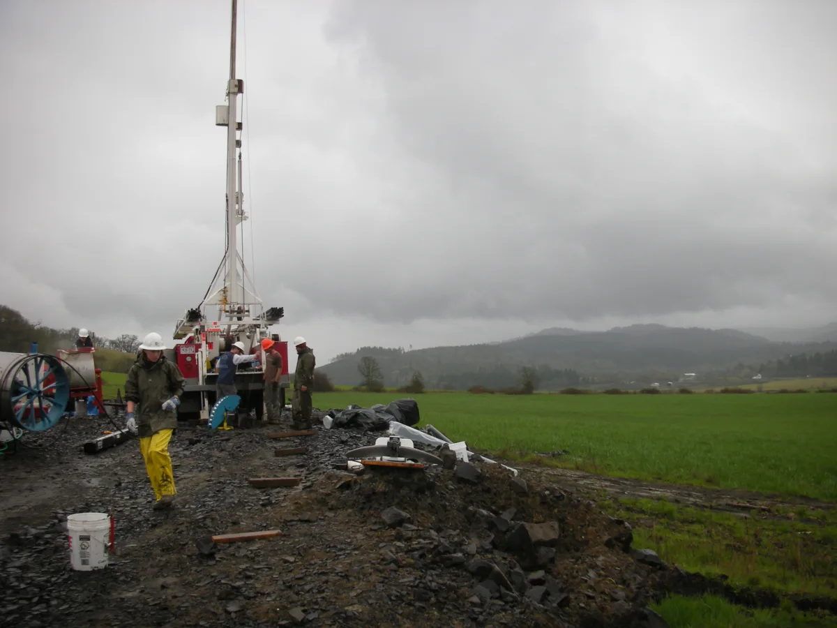 Workers surround a drill rig in a muddy patch surrounded by green grass.