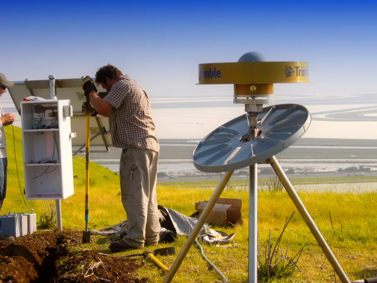 A worker digs a trench to connect the GPS antenna to the enclosure, with a large body of water in the background.
