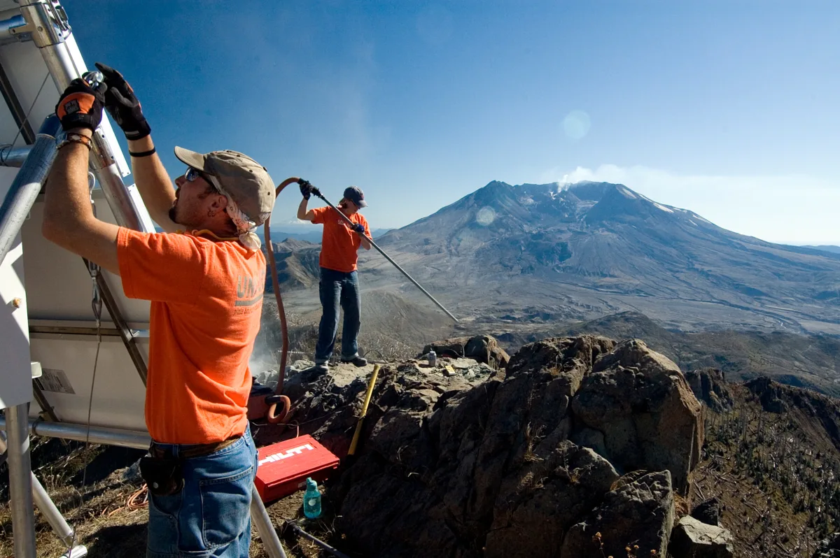 Two workers manipulate equipment on a rocky outcrop, with Mount St. Helens in the background.