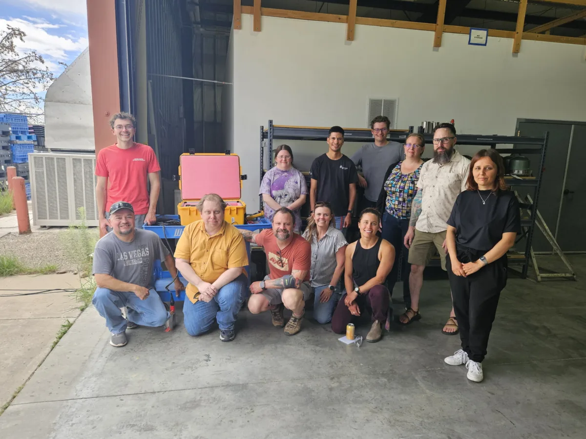 A group of twelve people pose for a photo inside a warehouse-like space, some standing and some kneeling in front. Behind them are industrial shelves and bright yellow and blue transport cases, with one case open to reveal pink foam padding.