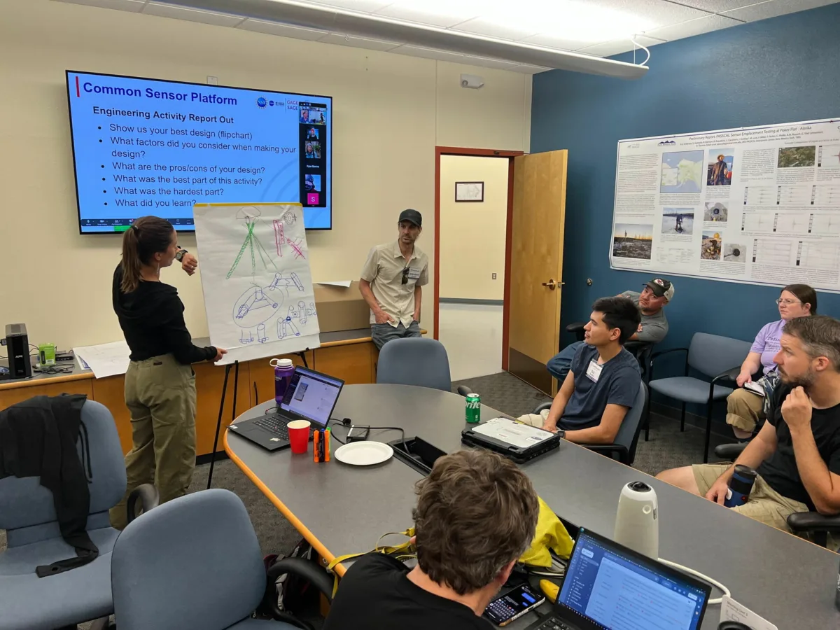 A woman stands at the front of a conference room, presenting a hand-drawn engineering diagram on a flip chart to a seated group. A man stands beside her, and a large screen behind them displays a slide titled “Common Sensor Platform: Engineering Activity Report Out,” with a list of discussion questions. Several participants watch attentively, with laptops, drinks, and notes on the table.