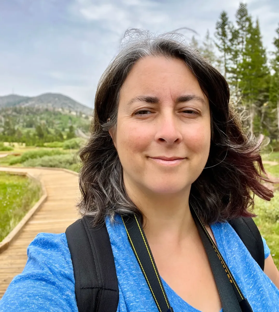 Photo of a person with should-length brown hair and a blue shirt wearing a backpack, with a path and hills in the background.