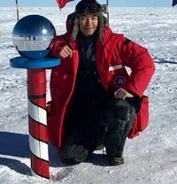 Photo of person kneeling near the South Pole marker wearing a red coat and fur cap.
