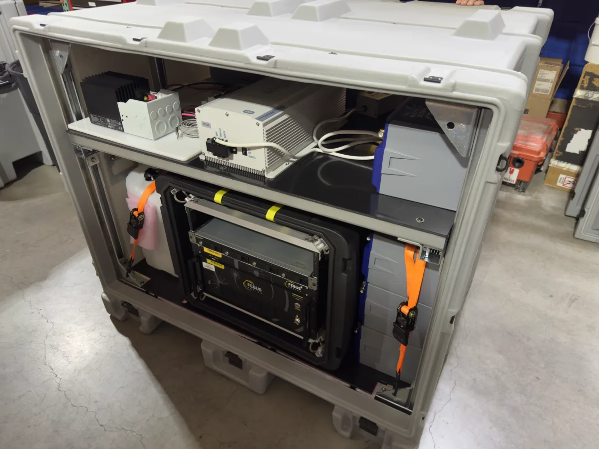 Open rugged gray equipment enclosure showing a neatly organized DAS power system. The upper shelf holds electronic control units and wiring, while the lower section contains a rack-mounted component and large battery modules secured with bright orange straps.
