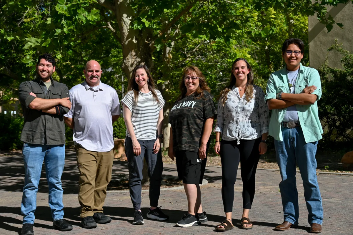 Six people in a row smile at the camera, framed by trees in the background.