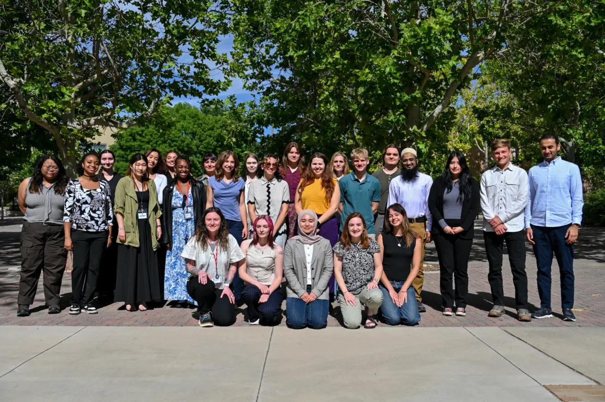 26 NSF NGF interns line up in three rows for a group picture. Five interns kneel in the front on concrete, while the other two rows are interspersed. They are framed by trees in the background.