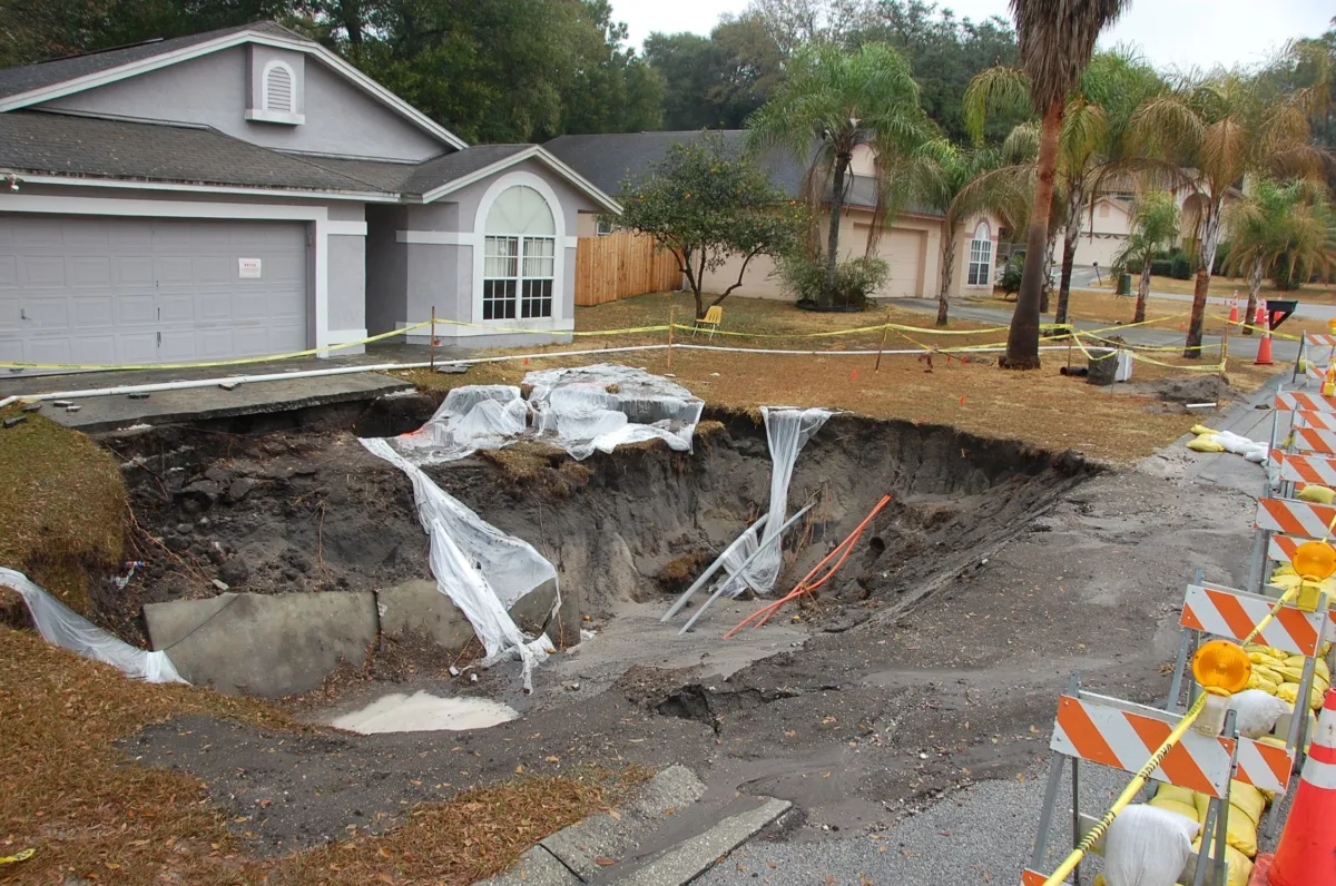 A suburban street lined with houses, with a large sinkhole in the front yard of the closest house. The sinkhole has been roped off with caution tape.