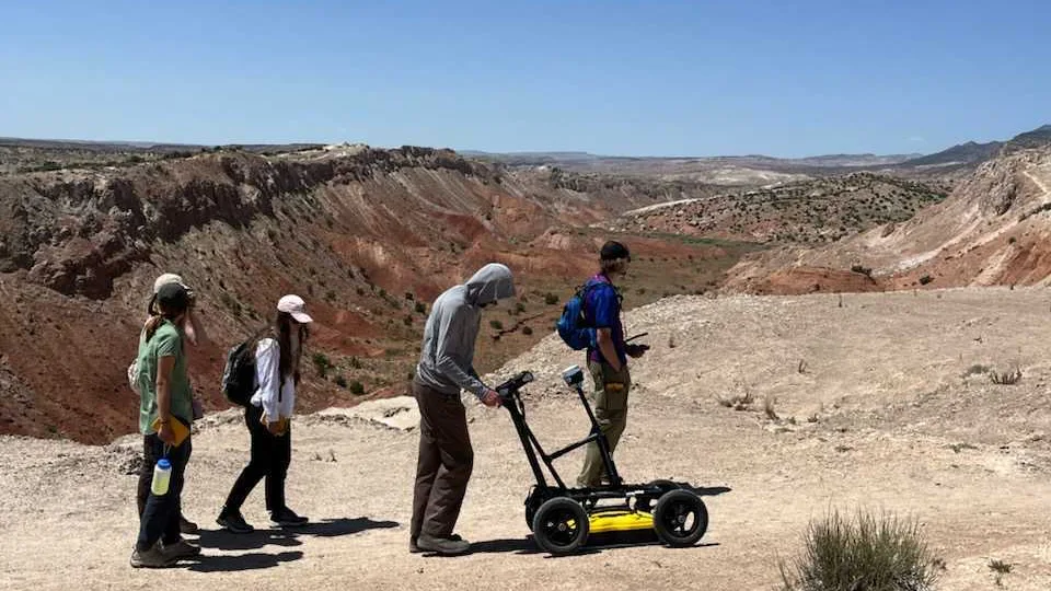 Five students walk on a flat, sandy cliff in front of desert mountains. One is pushing a lawnmower-style GPR system.