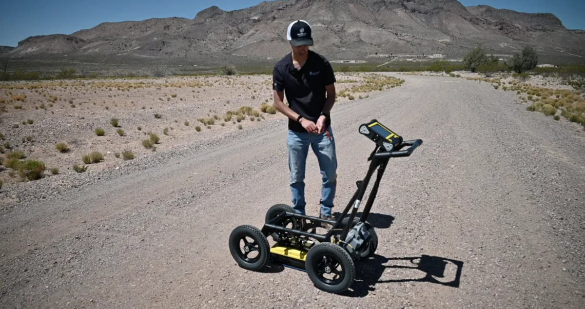 A man standing with a "lawnmower"-style GPR instrument in front of him on a gravel road. There are desert mountains in the background.