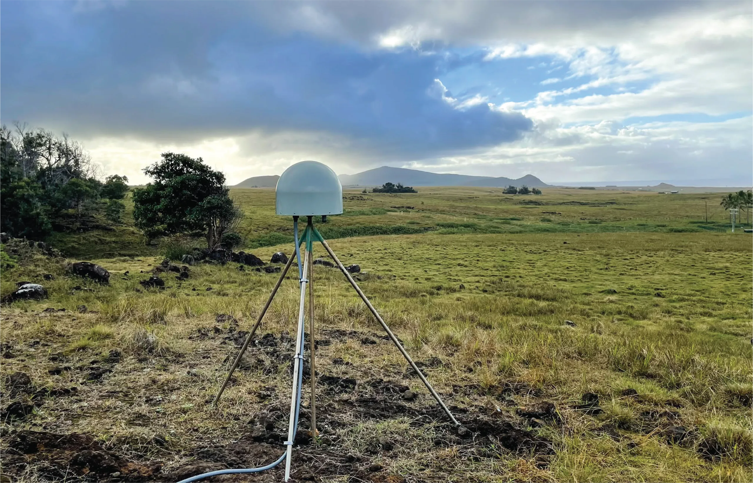 Photo of a permanent GNSS station in a grassy field with trees and clouds in the background.