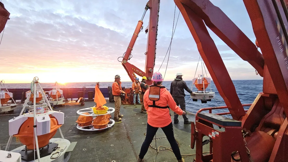 Researchers on a ship use a crane to lower an orange GNSS seafloor sensor into the ocean. There are additional sensors on the deck.