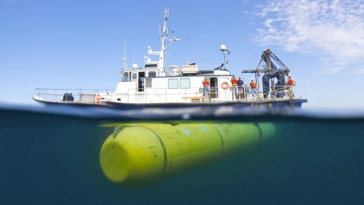 A split-level view of a research vessel above the water and a yellow Autonomous Underwater Vehicle (AUV) submerged beneath it.