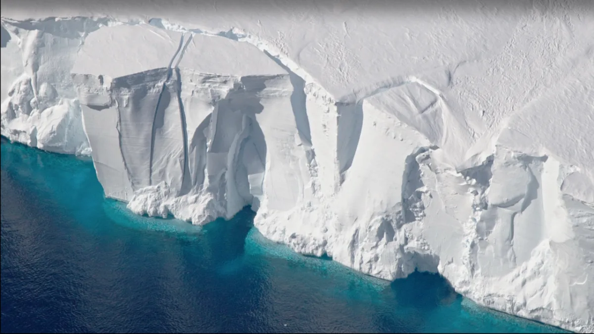 An image of the edge of the Antarctic Ice Sheet as it touches clear, blue water. A large chunk of the ice sheet appears to be breaking off.