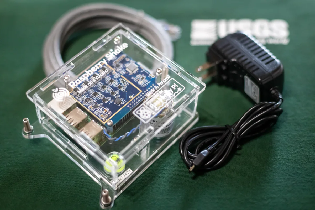 A close-up view of an unplugged Raspberry Shake seismometer sitting on green fabric with "USGS" in the background.