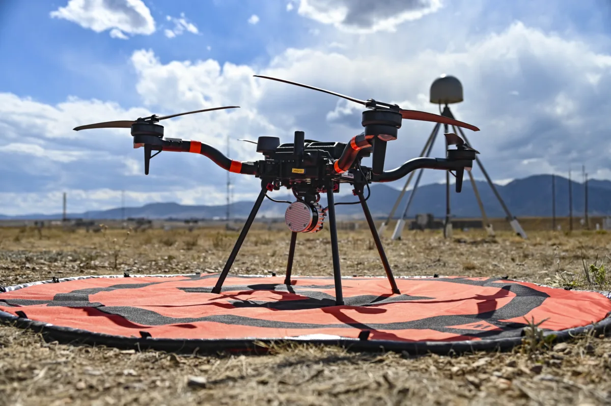 Close photo of a drone on the ground with a GPS station and mountains in the background.