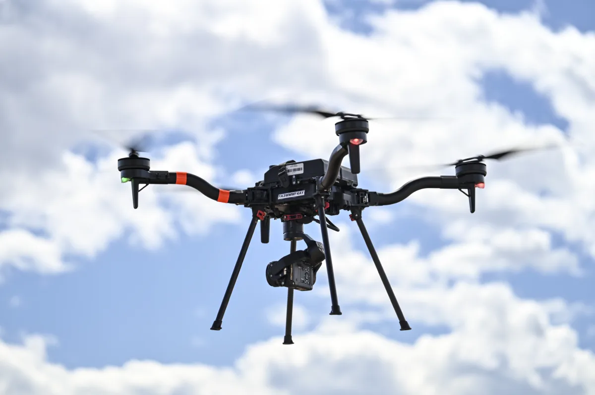 Drone in flight carrying a camera, with clouds behind.