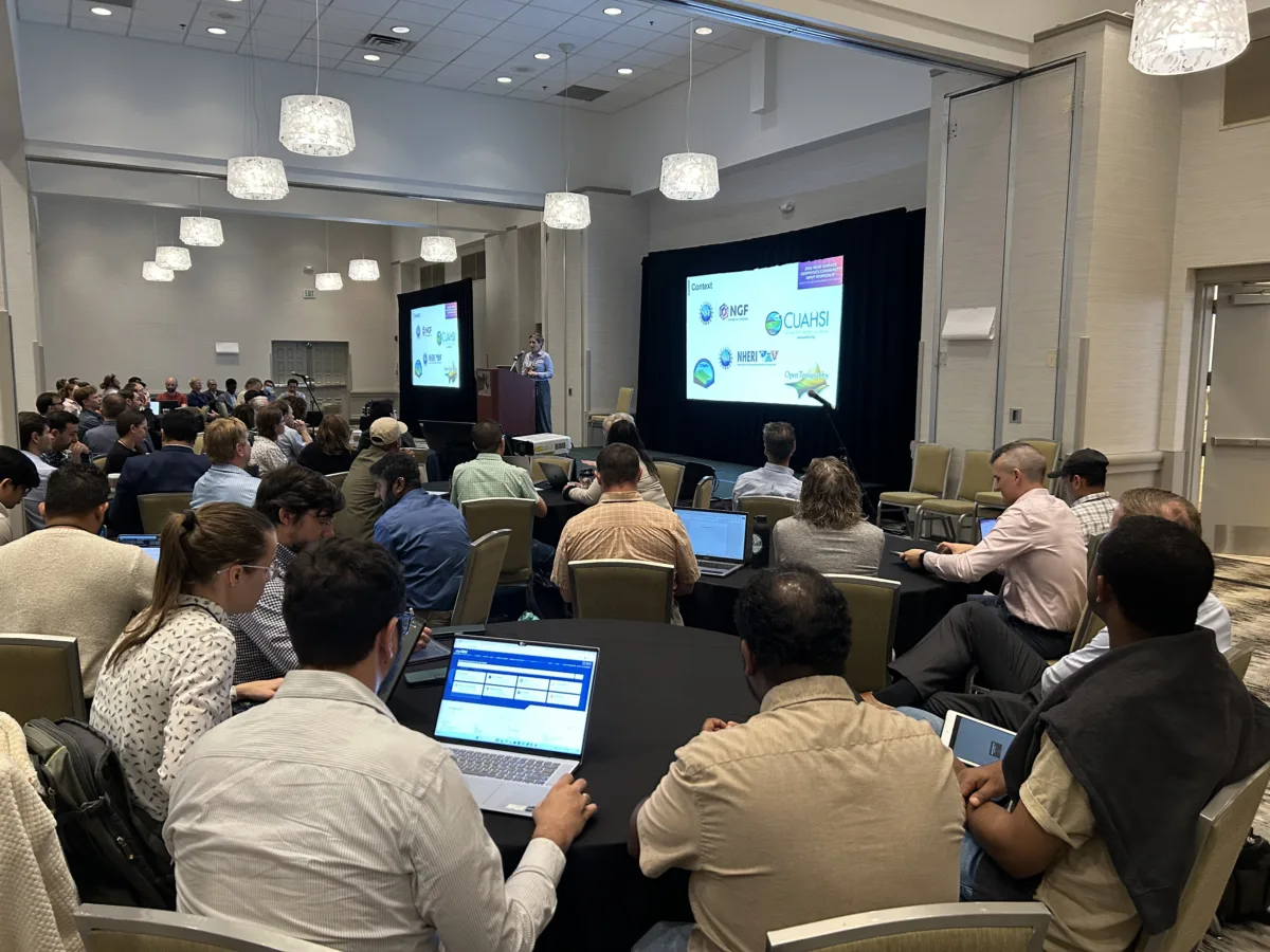 Attendees seated at round tables listen to a presenter speaking at a podium during a conference session. A large screen displays the logos of several geoscience organizations.