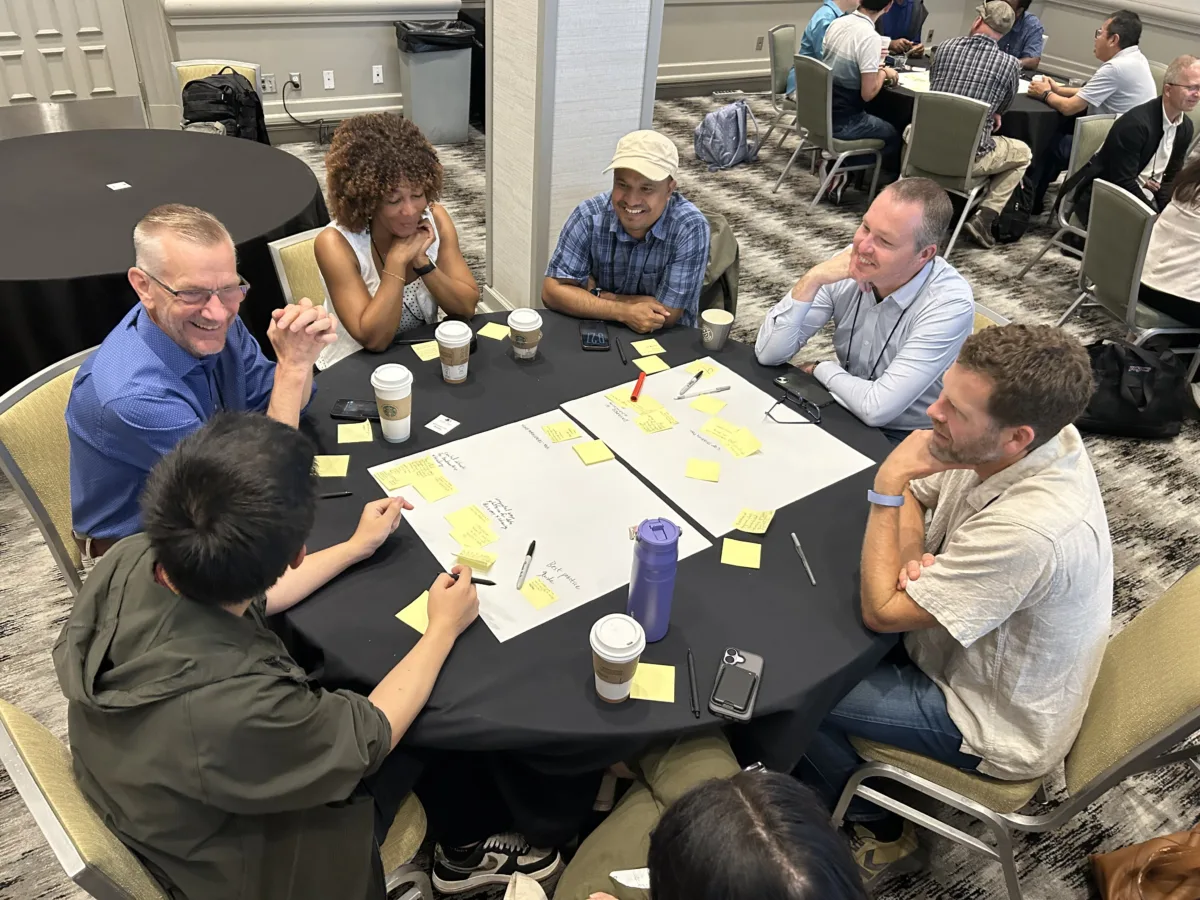 Six conference attendees participate in a small-group discussion around a table covered with sticky notes, markers, and large sheets of paper.